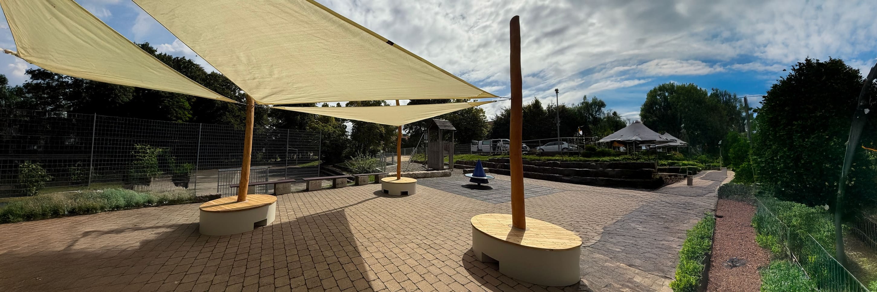 Terrassenbereich mit großen Sonnensegeln, runden Holzbänken, Ziegelsteinpflaster, Grünpflanzen und einem teilweise bewölkten Himmel.