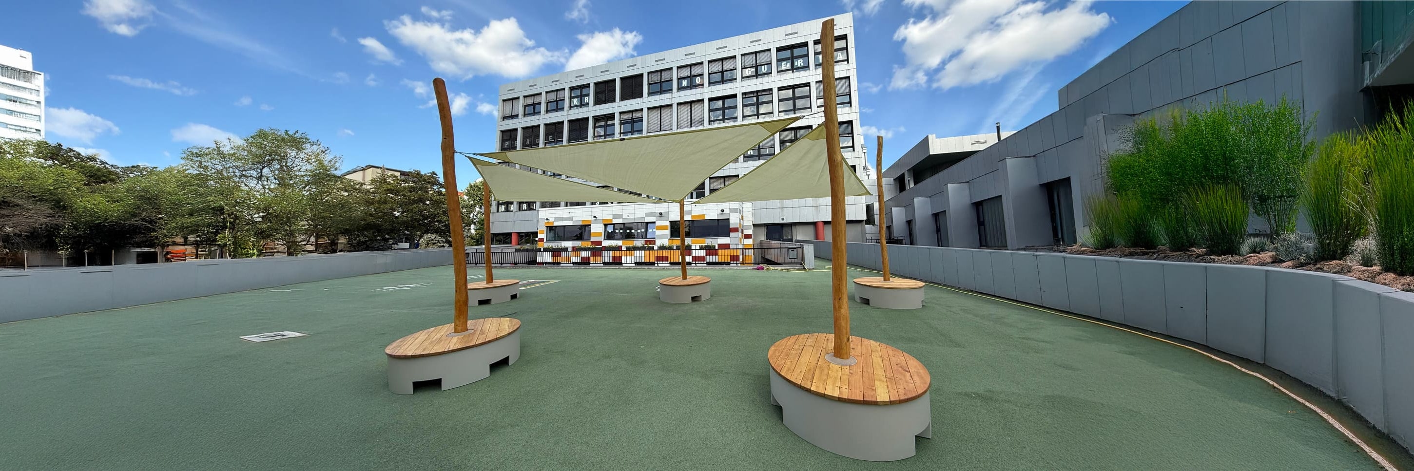 Dachterrasse mit grünem Bodenbelag, vier hölzerne Sitzgelegenheiten mit Sonnensegeln, moderne Gebäude, Bäume und ein blauer Himmel mit Wolken im Hintergrund.