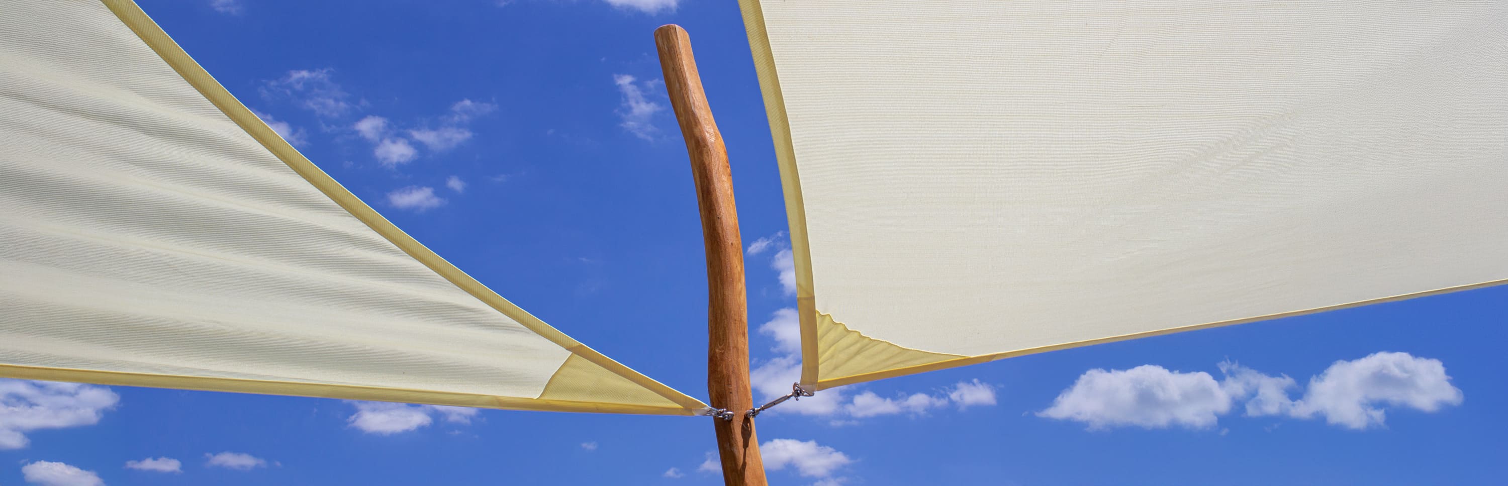 Zwei beigefarbene Sonnenschirme aus Stoff sind an einer Holzstange vor einem blauen Himmel mit vereinzelten weißen Wolken befestigt.