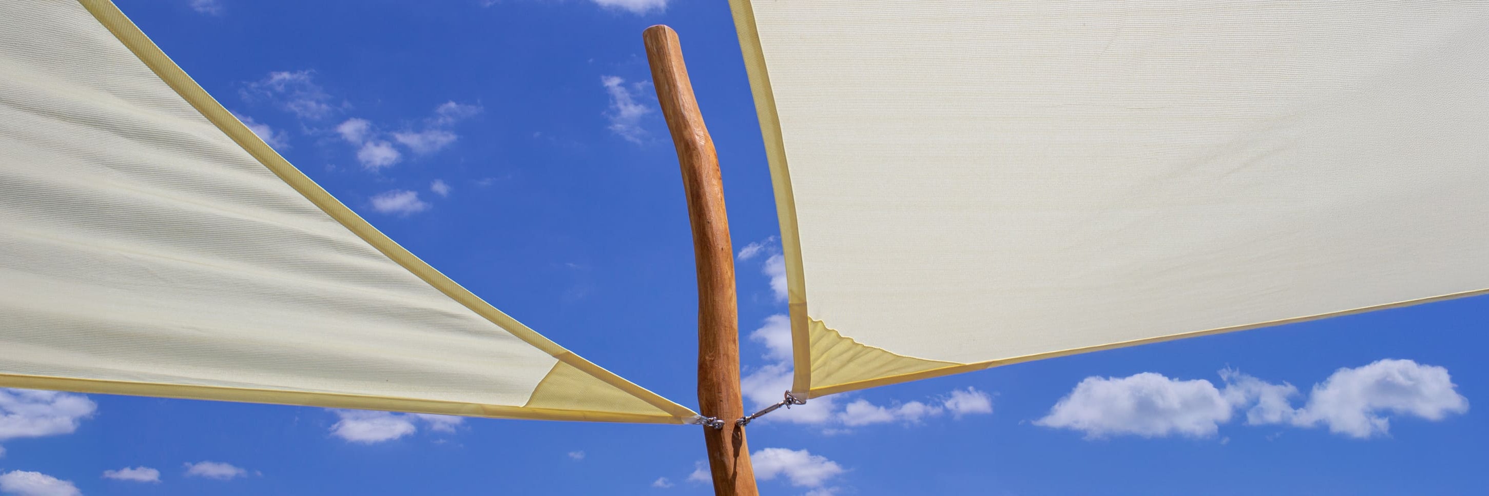 Zwei beigefarbene Sonnenschirme aus Stoff sind an einer Holzstange vor einem blauen Himmel mit vereinzelten weißen Wolken befestigt.