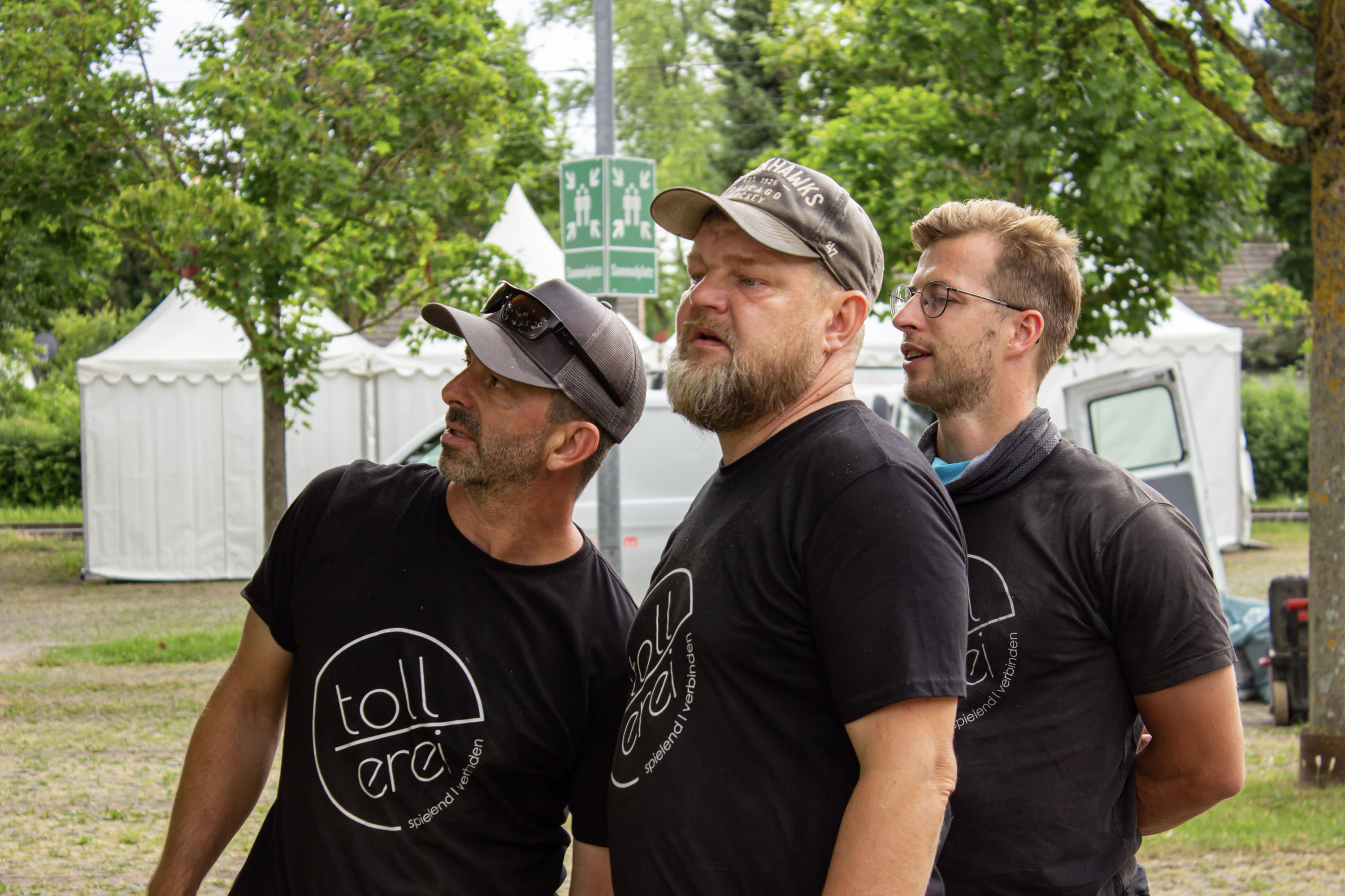 Drei Männer in passenden schwarzen tollerei-Tshirts stehen im Freien und schauen in die gleiche Richtung. Im Hintergrund sind weiße Zelte und Grünpflanzen zu sehen.
