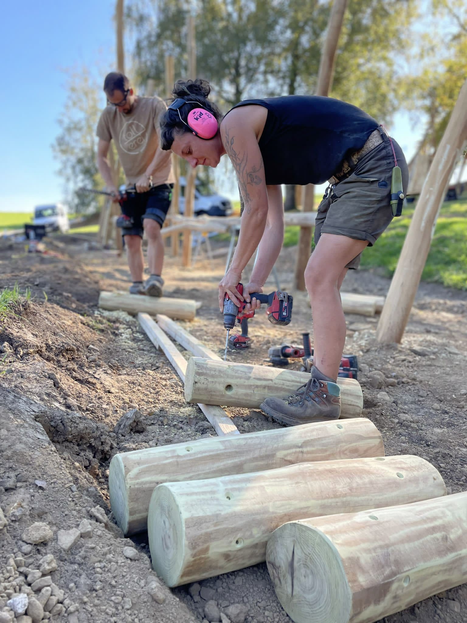 Zwei Personen arbeiten im Freien an einem Bauprojekt und befestigen mit einer Bohrmaschine große Holzstämme im Boden. Sie tragen Schutzkleidung.