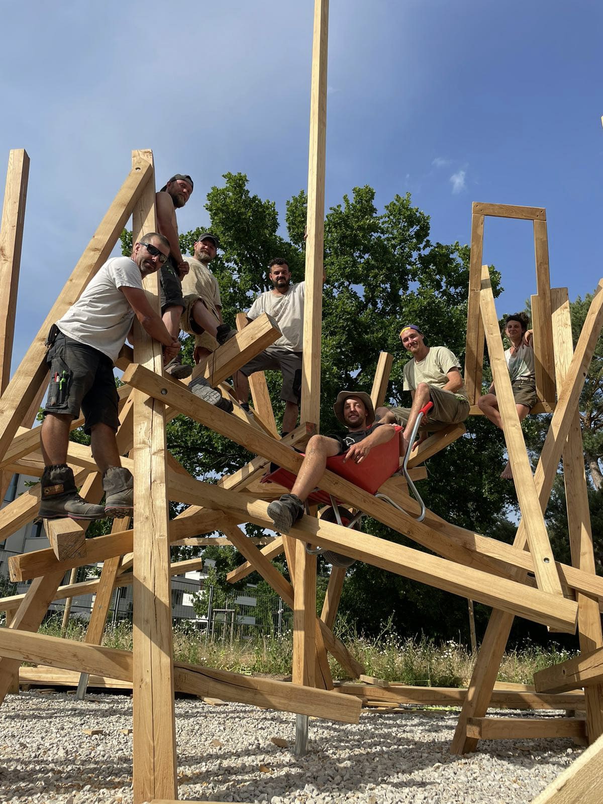 Eine Gruppe von Menschen in Freizeitkleidung posiert auf einer großen geometrischen Holzklettergerüst, mit Bäumen und Gras im Hintergrund.