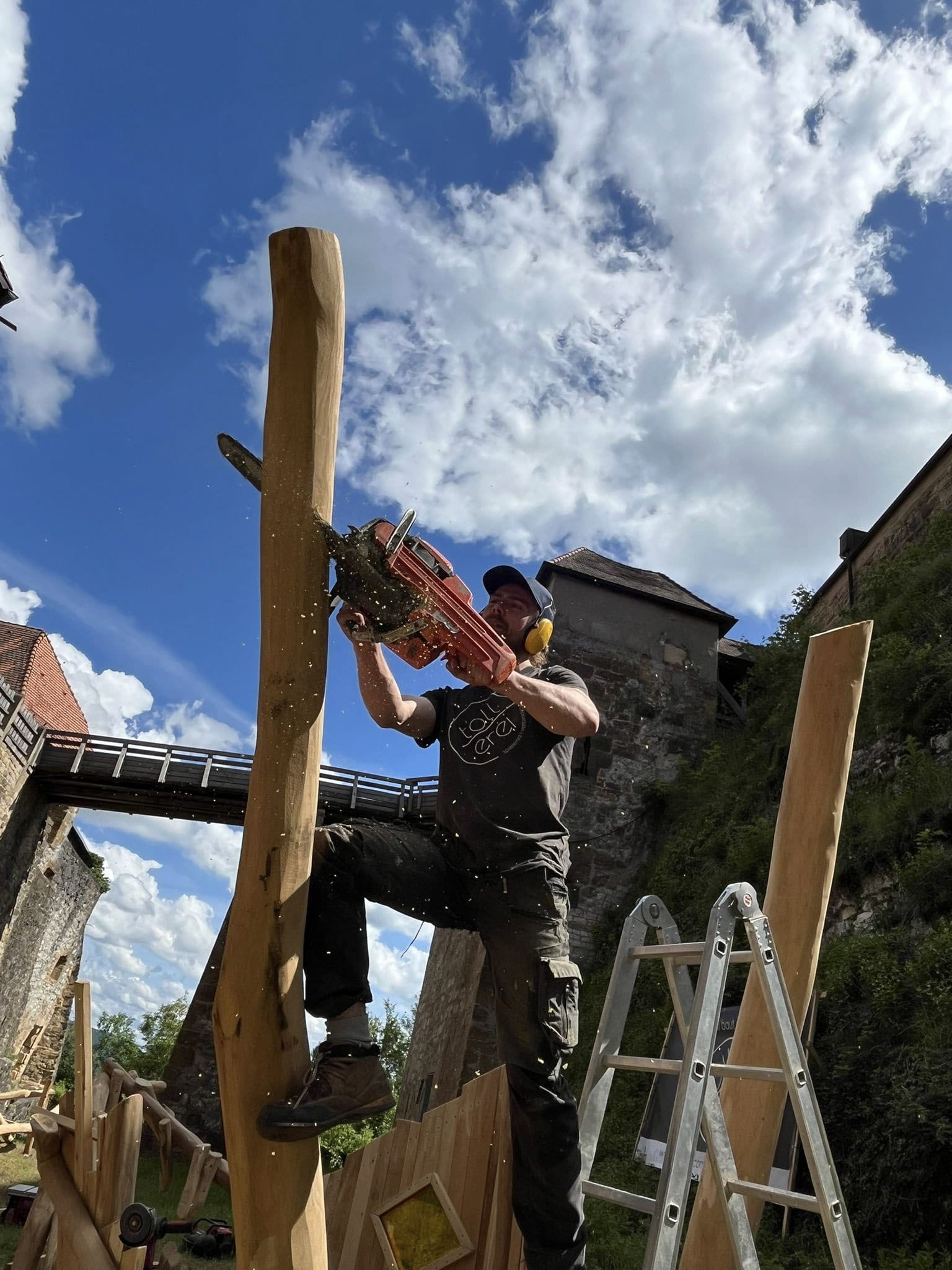Eine Person mit Gehörschutz sägt mit einer Kettensäge ein hohes Holzbrett im Freien, mit einer Leiter in der Nähe und Gebäuden im Hintergrund unter einem teilweise bewölkten Himmel.