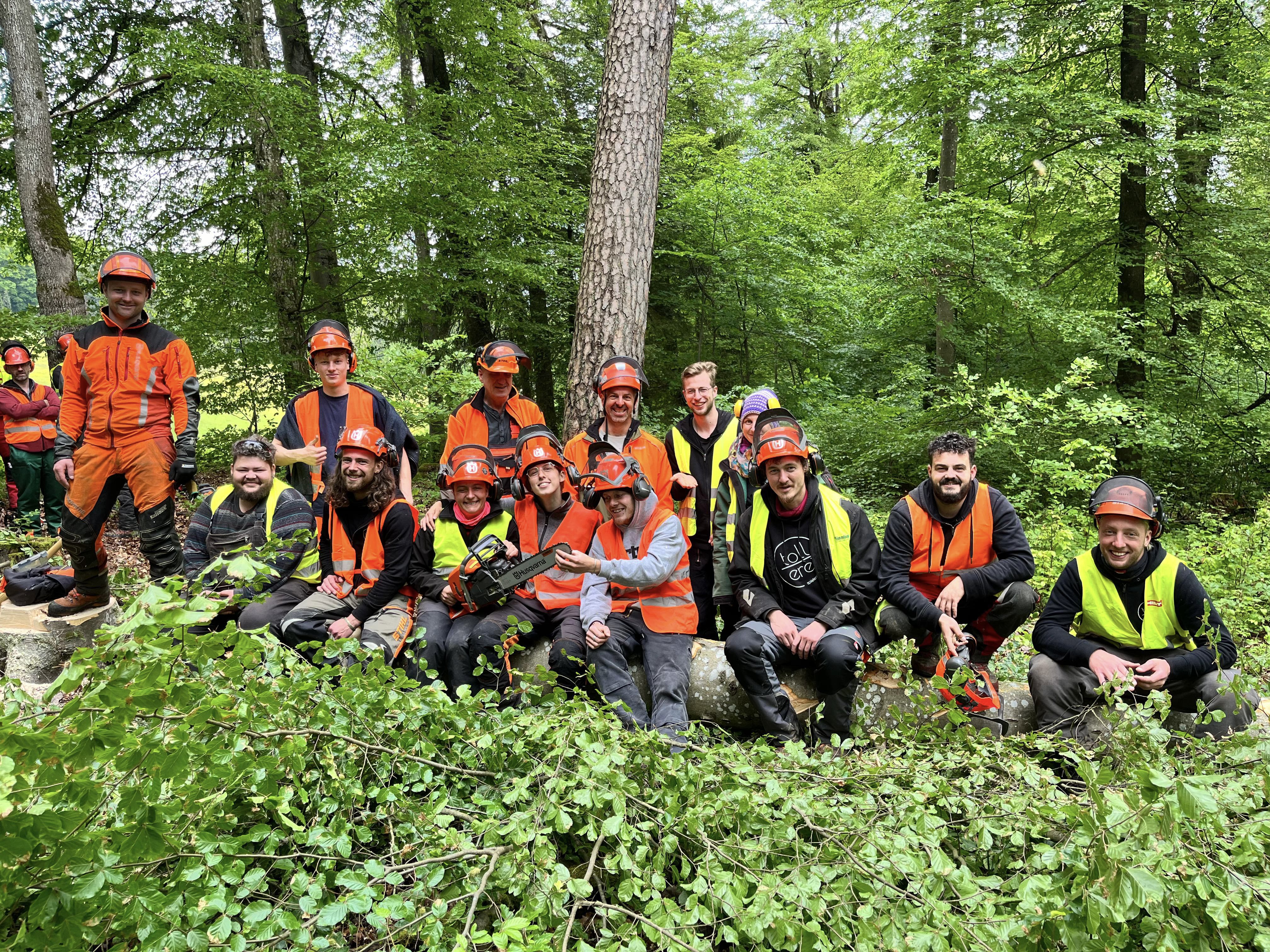 Eine Gruppe von Menschen in Schutzausrüstung und mit Helmen posiert in einem grünen Wald, sitzend und stehend hinter abgeschnittenen Ästen und Baumstämmen.