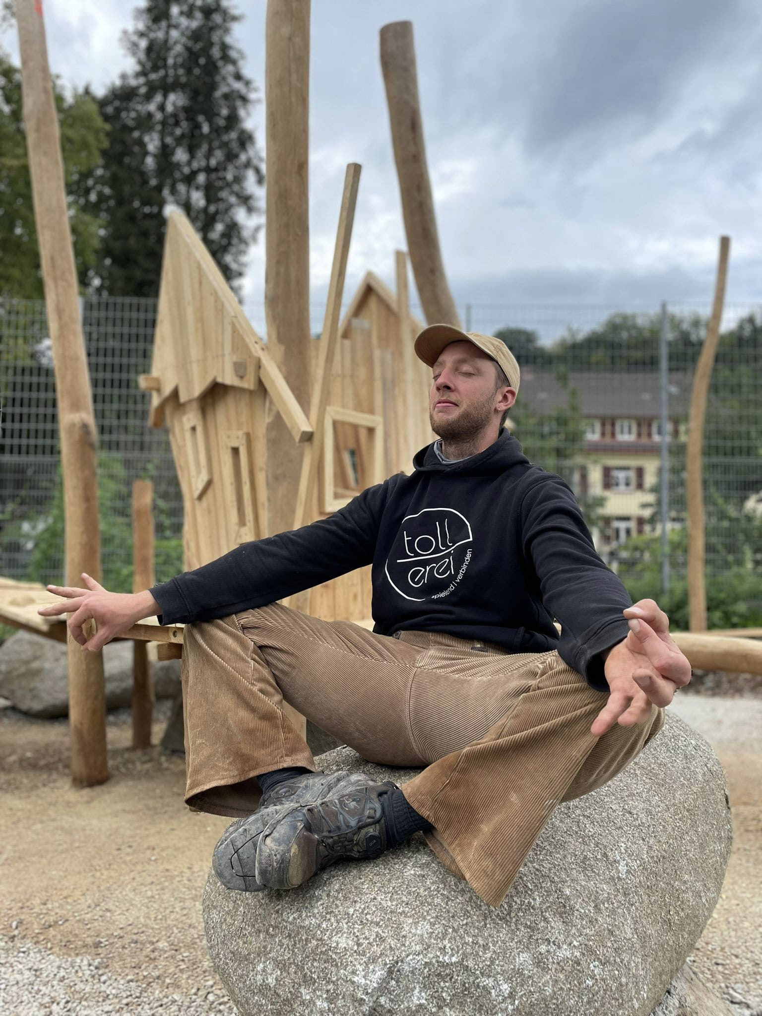 Ein Mann in legerer Kleidung sitzt im Schneidersitz auf einem großen Felsen, die Augen geschlossen und die Hände in meditativer Haltung, mit Holzstrukturen und Bäumen im Hintergrund.