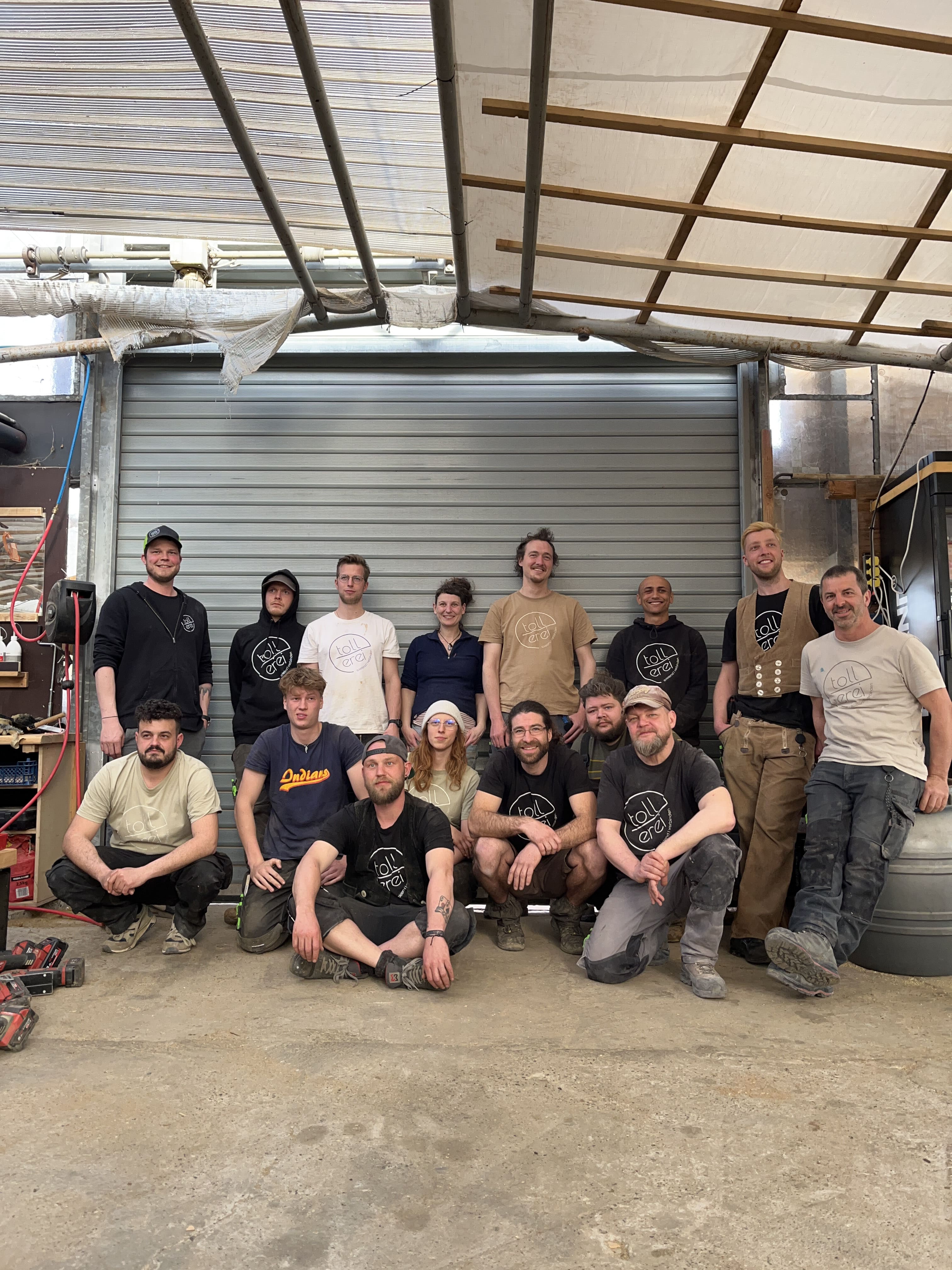 Eine Gruppe von fünfzehn Personen posiert zusammen in einer industriellen Werkstatt, mit Werkzeugen und Geräten um sie herum und einem Metalltor im Hintergrund.