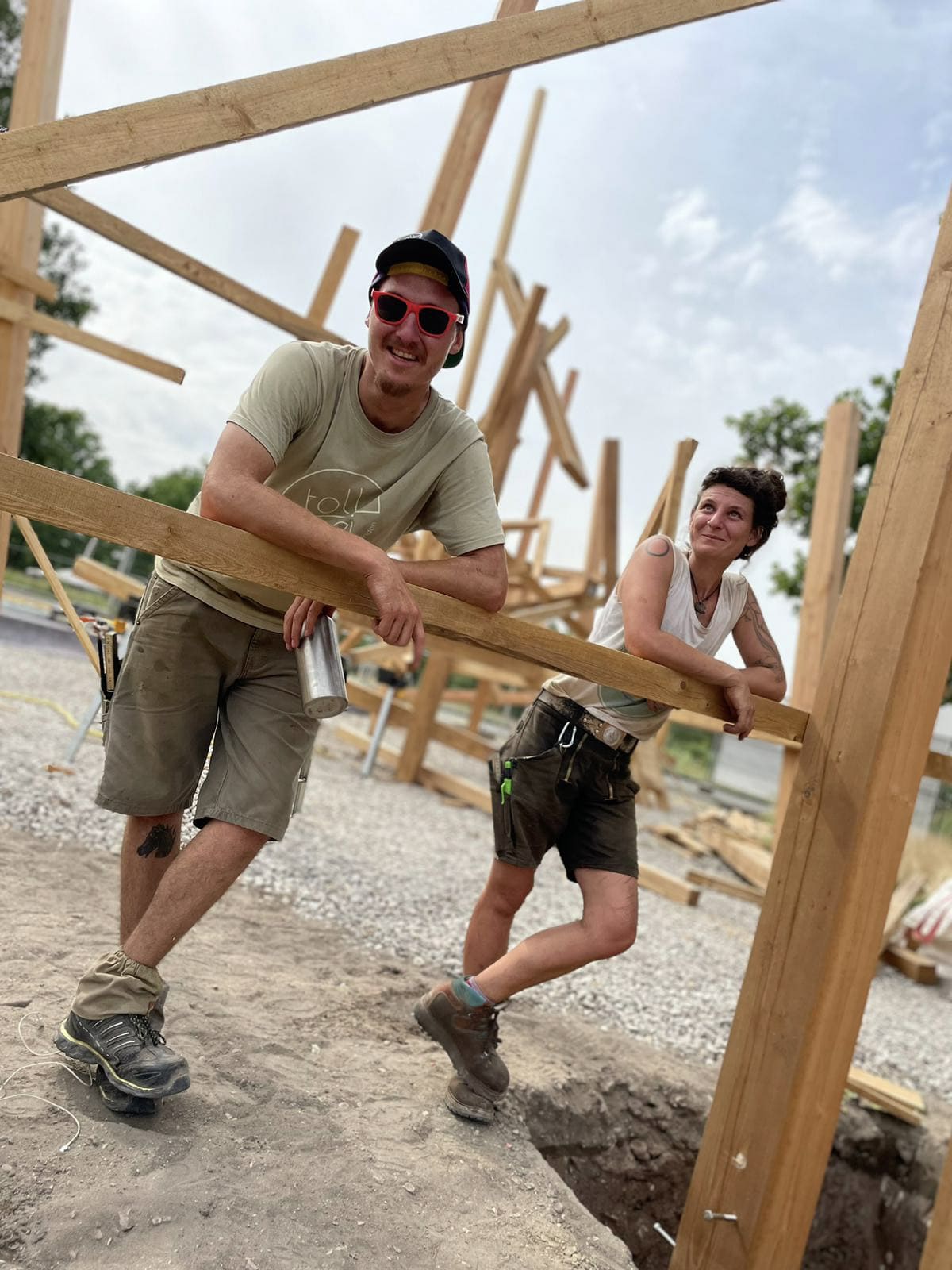 Zwei Personen in Arbeitskleidung und mit Werkzeuggürteln stehen auf einer Baustelle und lehnen sich an ein Holzgerüst, das von unfertigen Holzbalken umgeben ist.