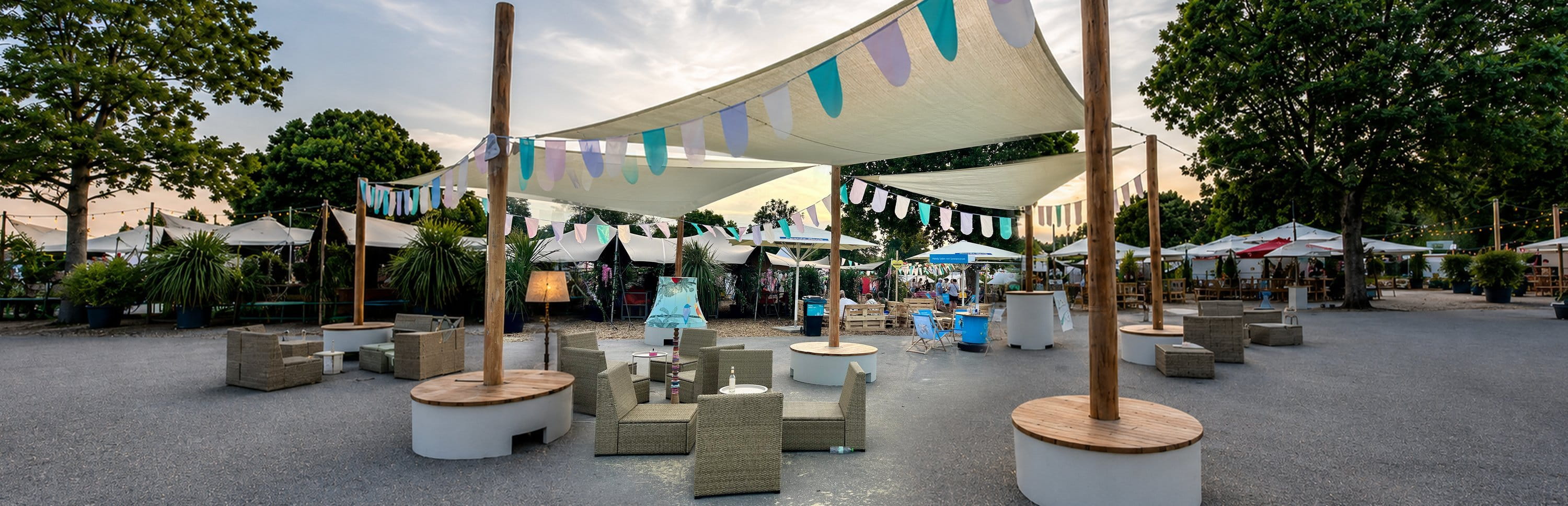Veranstaltungsfläche mit POP-UP-Sonnensegeln, bunten Wimpelketten, Loungemöbeln und runden Sitzpodesten am Abend.