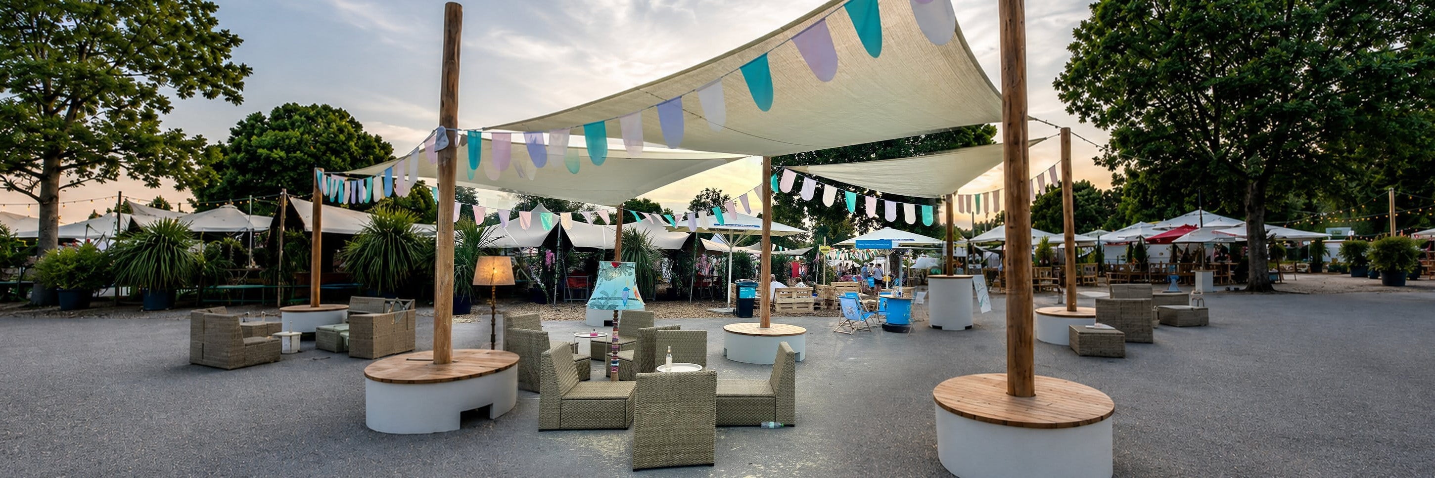 Veranstaltungsfläche mit POP-UP-Sonnensegeln, bunten Wimpelketten, Loungemöbeln und runden Sitzpodesten am Abend.