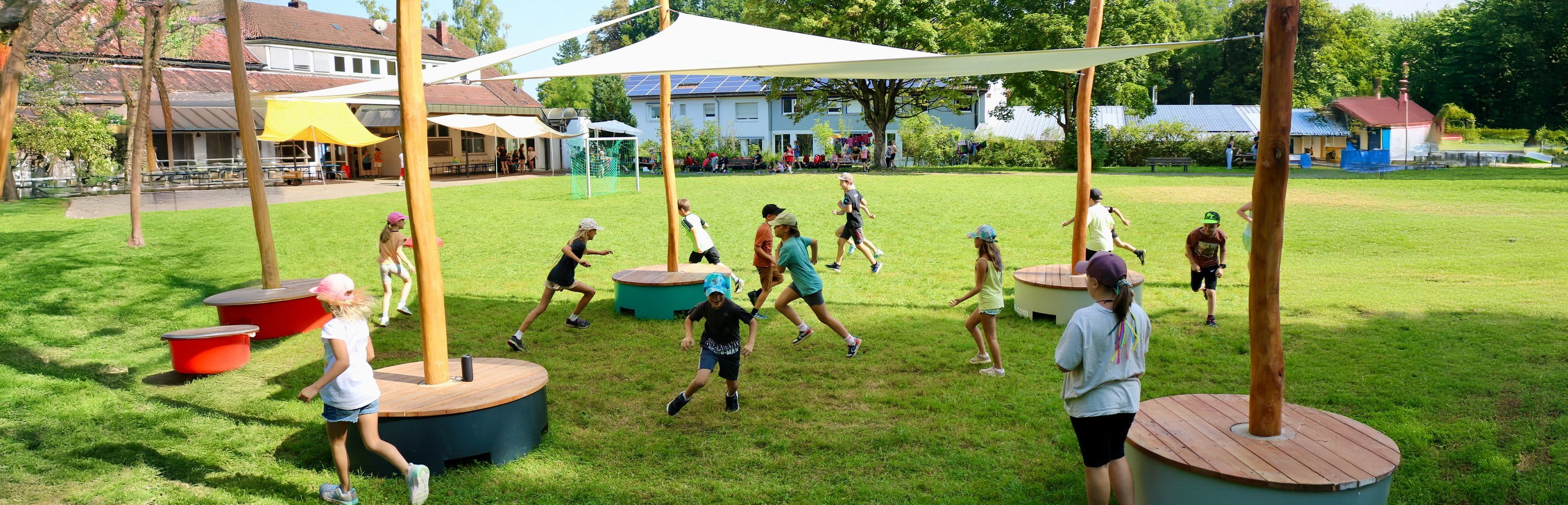 Kinder rennen und spielen auf einer grünen Wiese unter POP-UP-Sonnensegeln mit bunten Sitzpodesten.