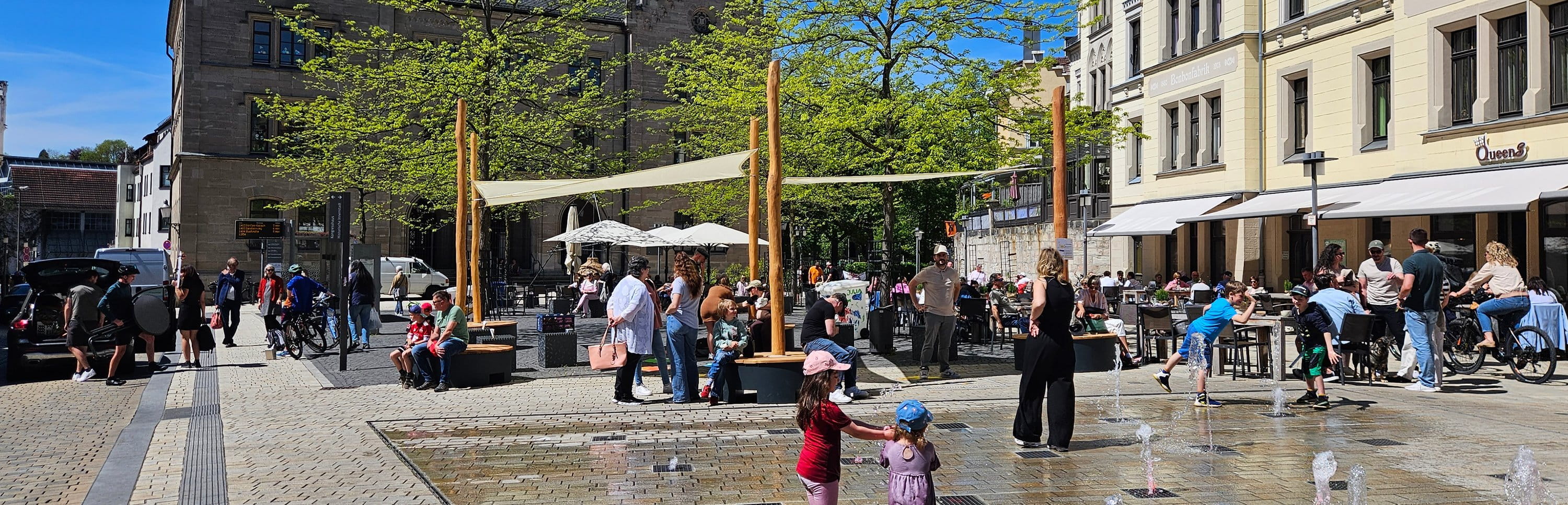 Belebter Innenstadtplatz mit POP-UP-Sonnensegeln über Sitzpodesten und Wasserfontänen, an denen Kinder spielen.