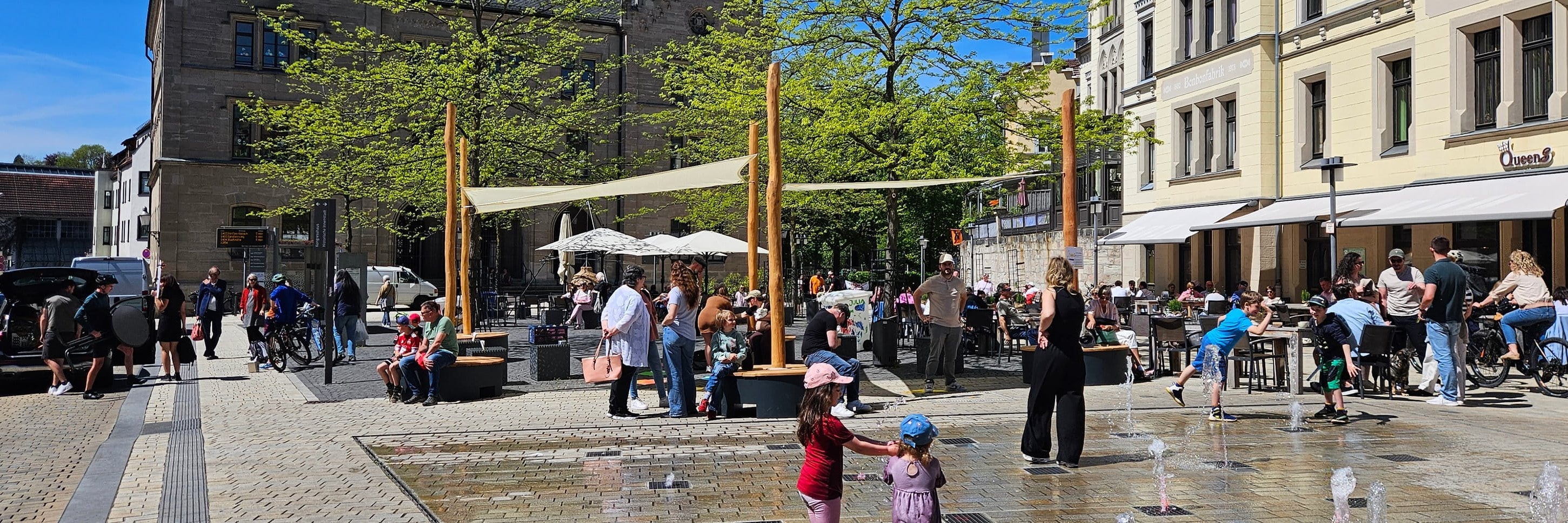 Belebter Innenstadtplatz mit POP-UP-Sonnensegeln über Sitzpodesten und Wasserfontänen, an denen Kinder spielen.