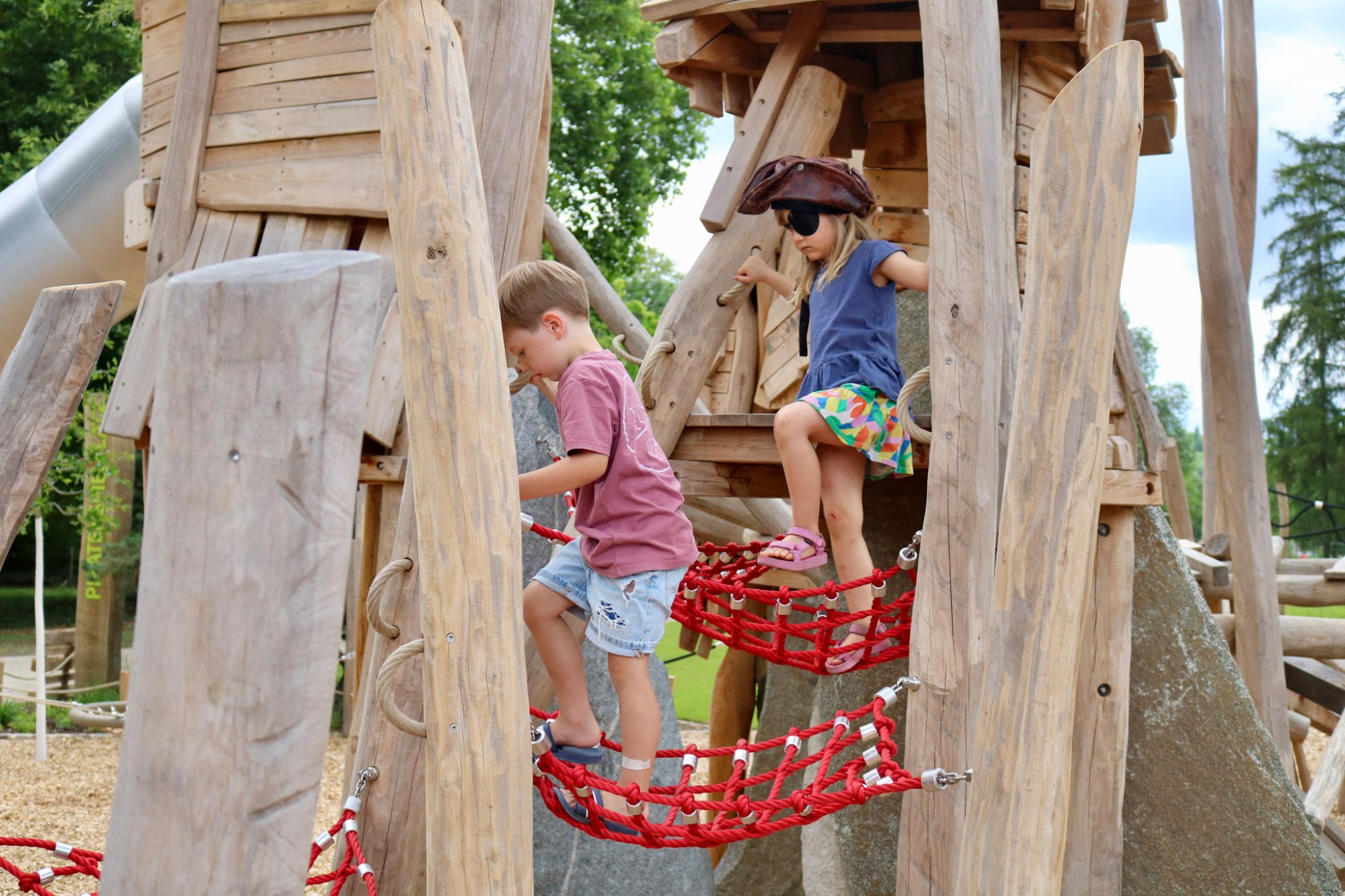 Zwei Kinder, eines mit Piratenhut, balancieren über eine rote Netzbrücke zwischen den Holztürmen des Piratenspielplatzes in Tübingen.