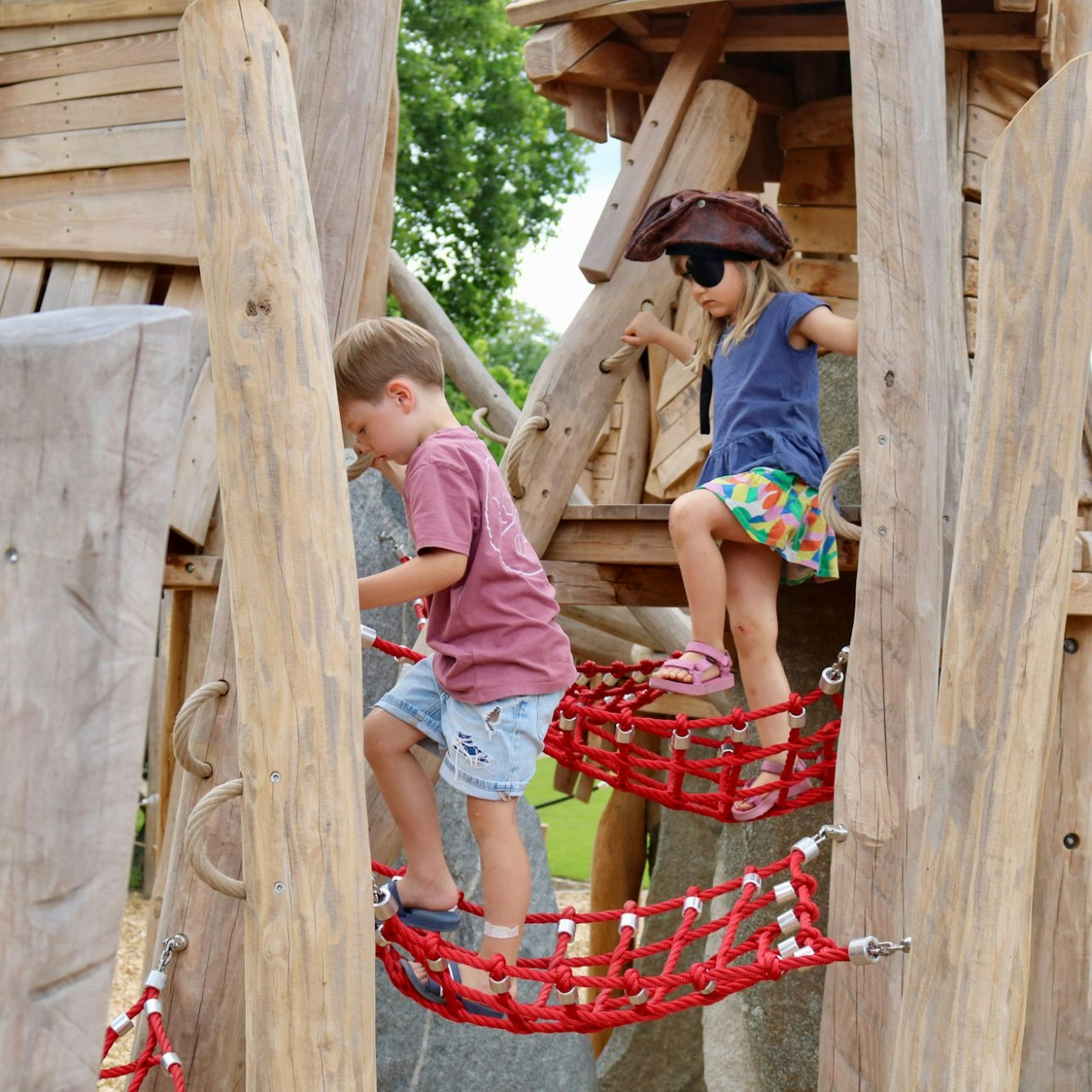 Zwei Kinder, eines mit Piratenhut, balancieren über eine rote Netzbrücke zwischen den Holztürmen des Piratenspielplatzes in Tübingen.