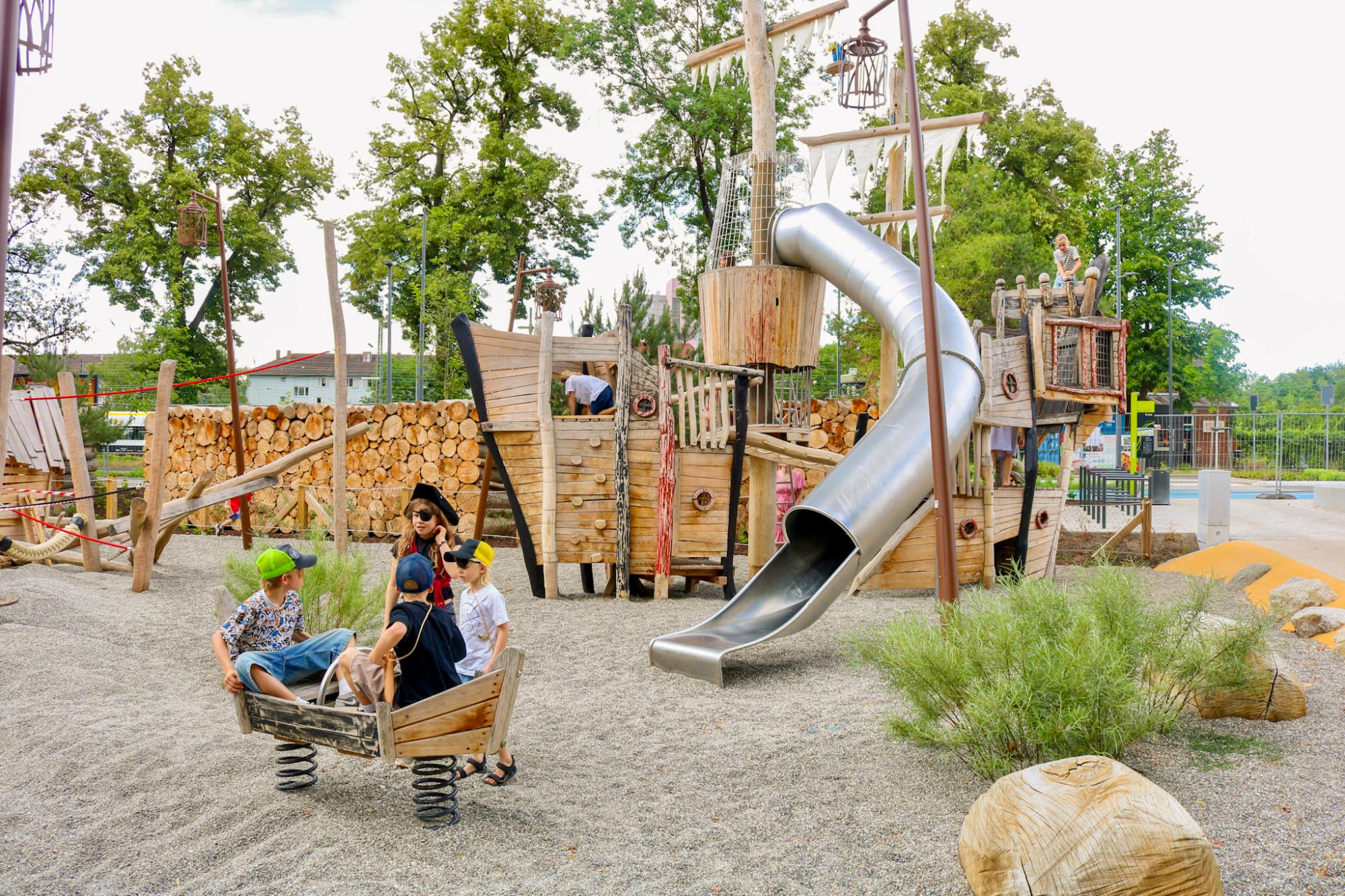 Übersicht des Piratenspielplatzes in Tübingen mit Holzschiff, Metallrutsche und Kindern in einem Federwipp-Boot im Sand.