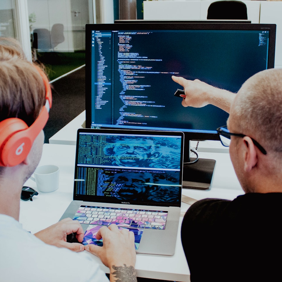En utvecklare sitter med händerna vilandes mot datorns mus, medan projektledaren pekar på den större externa skärmen.