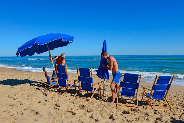 imagen de una playa con dos personas instalando material de playa (sombrillas y tumbonas).