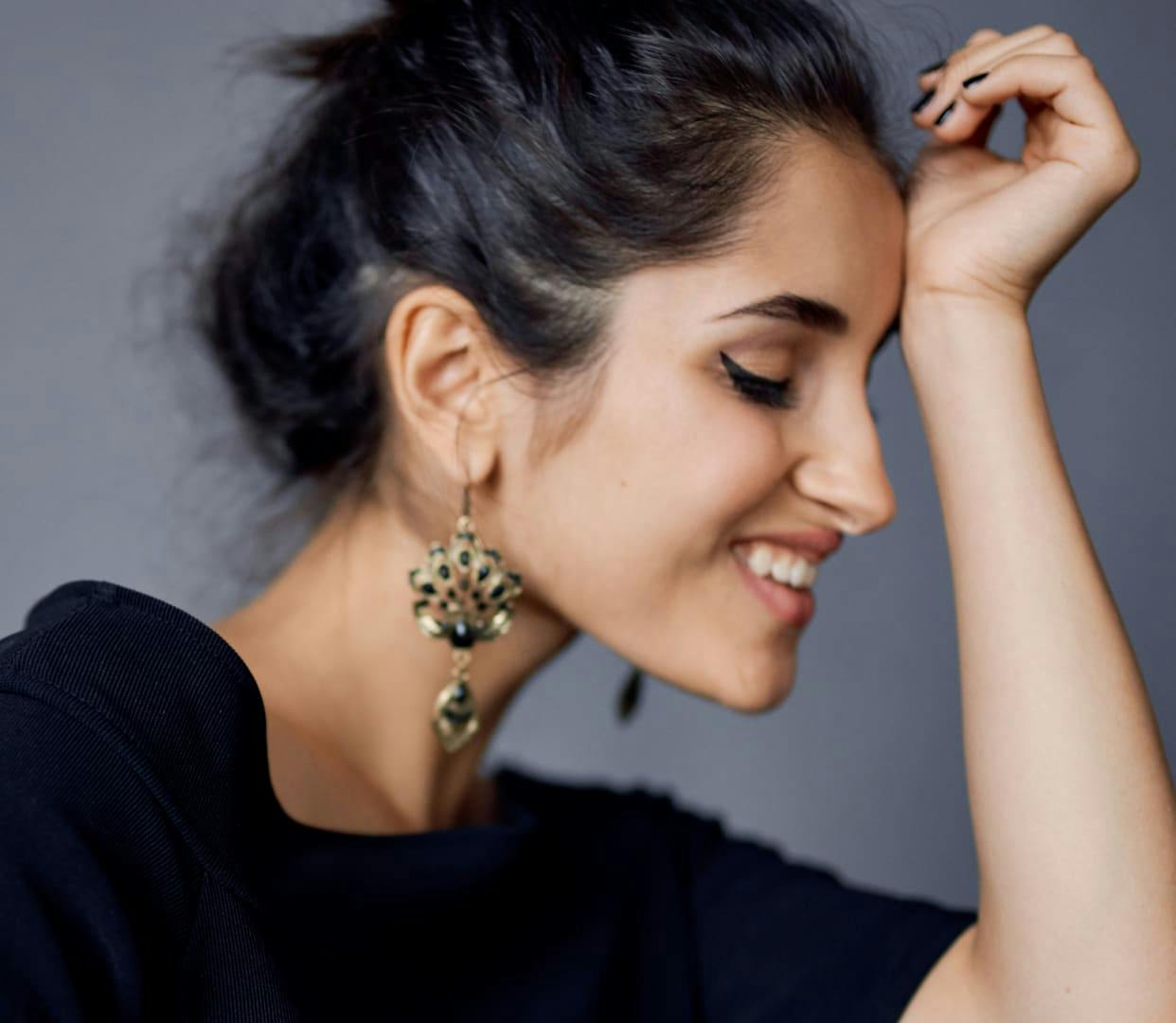 Woman with thick eyeliner and big earrings facing sideways smiling with her eyes closed and hand on her forehead