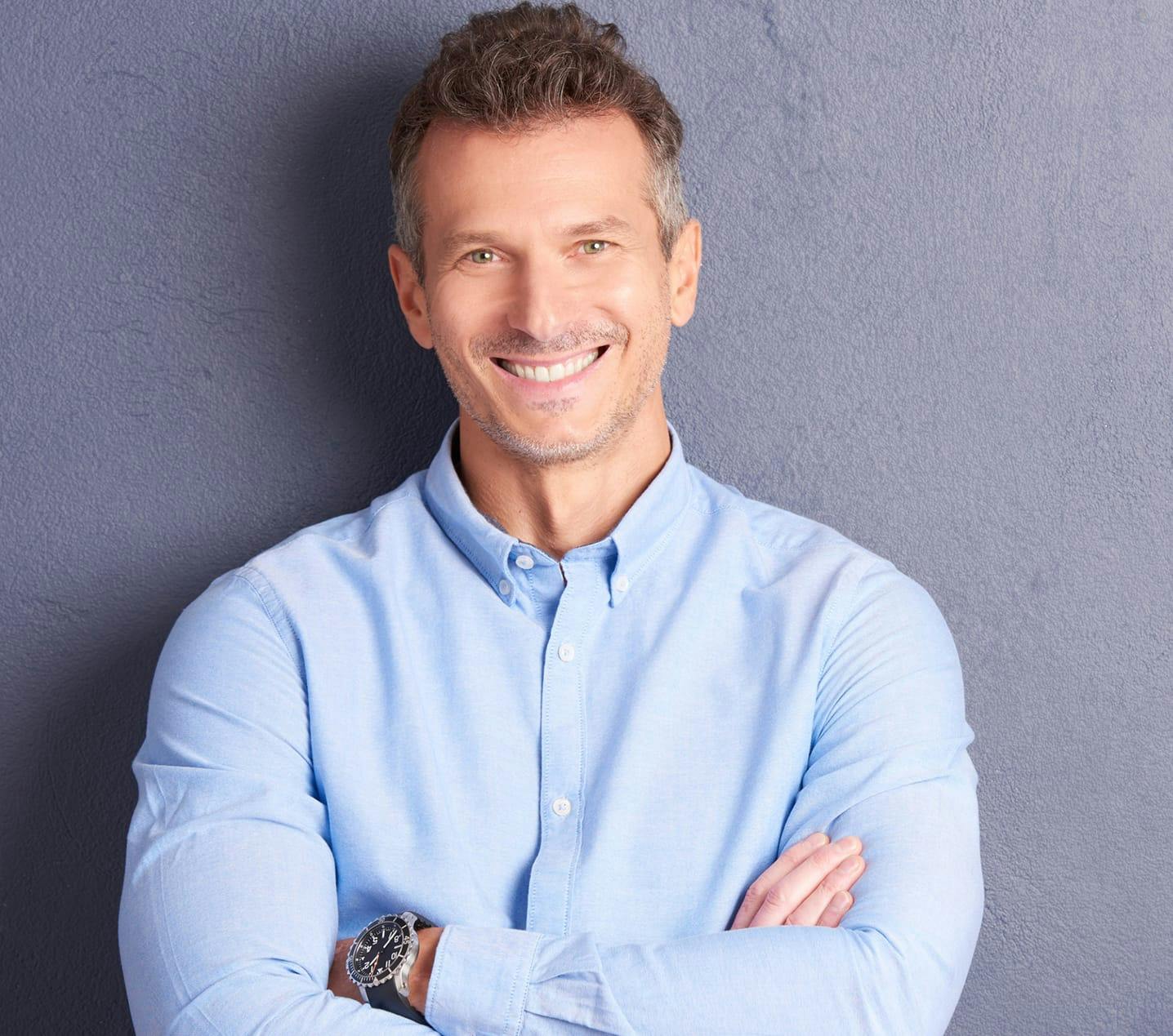 smiling man in blue shirt with arms crossed against a wall