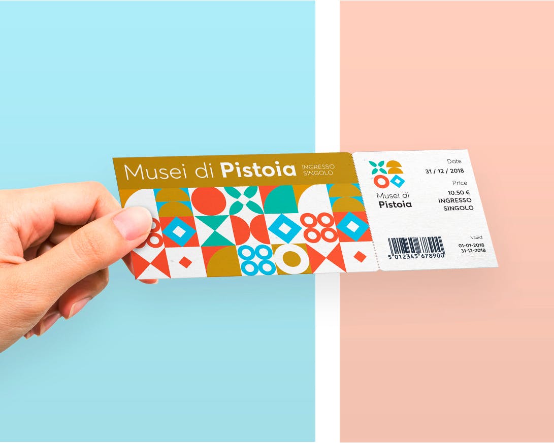 Una mano tiene un biglietto dei Musei di Pistoia con un design colorato su sfondo diviso tra azzurro e rosa, evidenziando arte e cultura.
