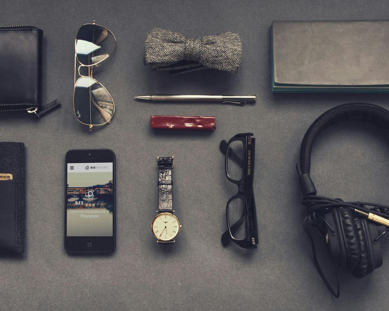 Una collezione di accessori maschili eleganti disposti su una superficie grigia, tra cui un portafoglio nero, un paio di occhiali da sole, un papillon grigio, una penna, un accendino rosso, uno smartphone, un orologio da polso, un paio di occhiali da vista, una cuffia audio e un taccuino.