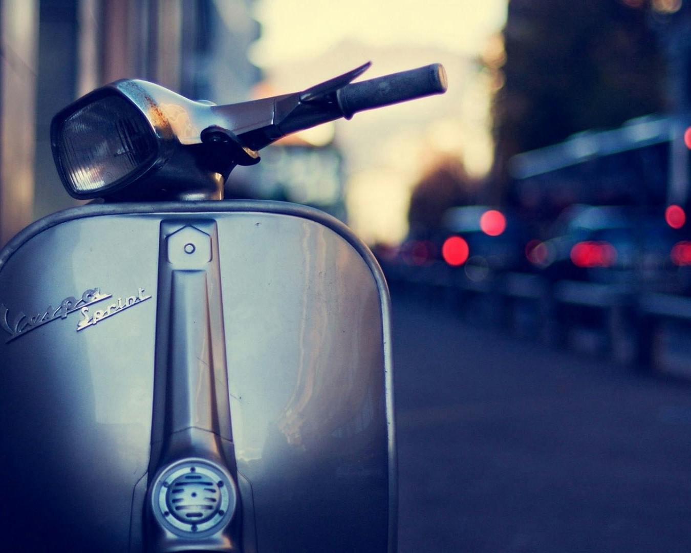 Una Vespa d'epoca argentata è parcheggiata su una strada cittadina al tramonto, con luci sfocate di traffico sullo sfondo.