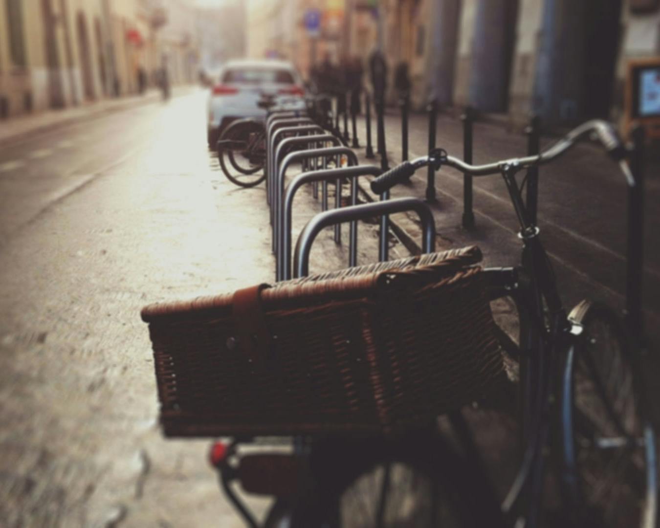 Strada cittadina al tramonto con una fila di biciclette parcheggiate; in evidenza una bicicletta con un cestino, mentre auto sono parcheggiate sullo sfondo.