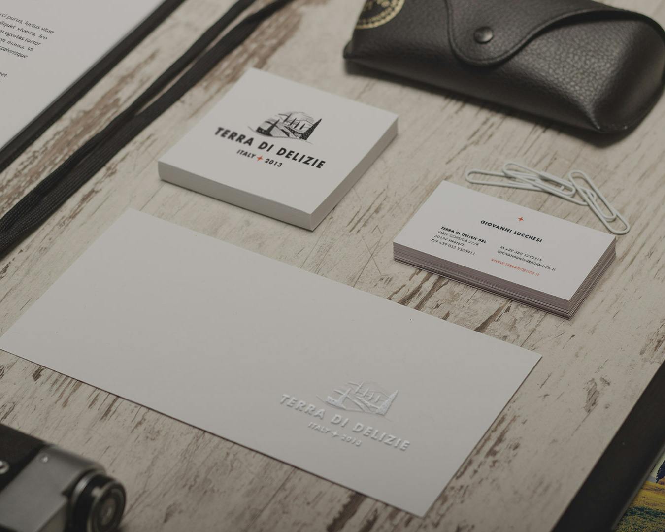 L'immagine mostra una collezione di articoli da ufficio su una scrivania in legno chiaro, tra cui biglietti da visita, una busta bianca con il logo "Terra di Delizie", una custodia per occhiali nera, una macchina fotografica e un cinturino in pelle nera, accompagnati da una stampa che ritrae un paesaggio.