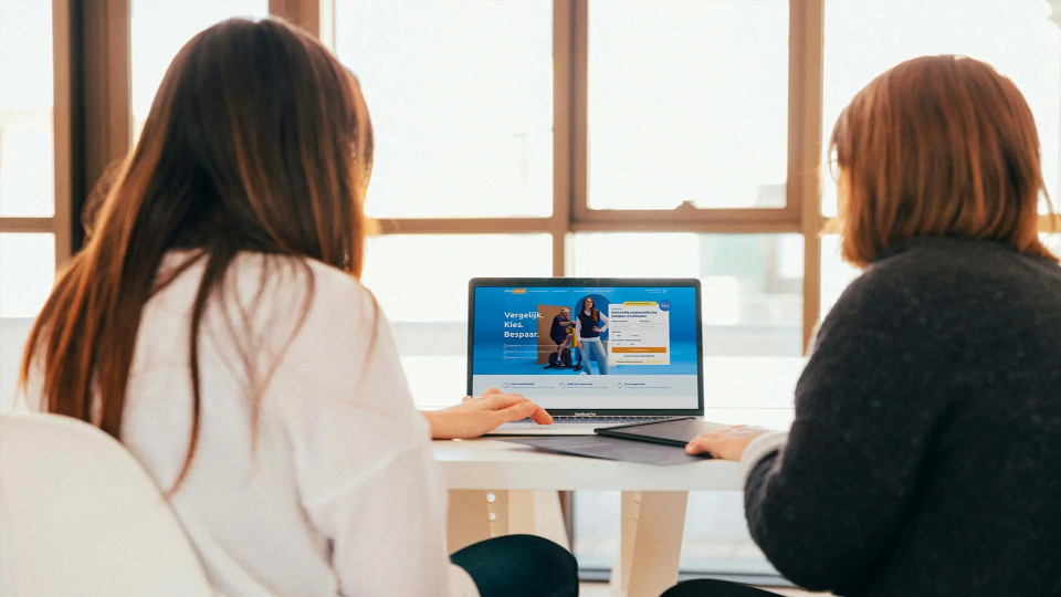 Twee vrouwen zitten aan een tafel en kijken naar een laptop met daarop een Zorgkiezer website. Ze kiezen een zorgverzekering. Ze zitten in een lichte kamer met grote ramen.