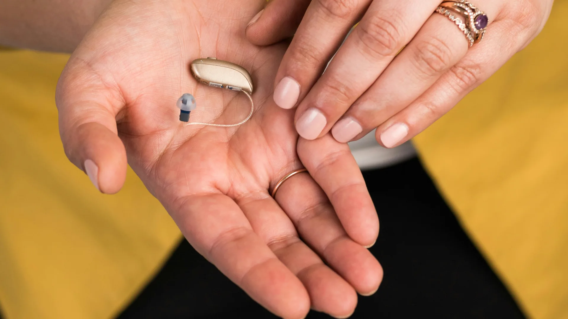 Close-up van uitgestrekte hand met gehoorapparaat tegen een achtergrond van gele kleding.