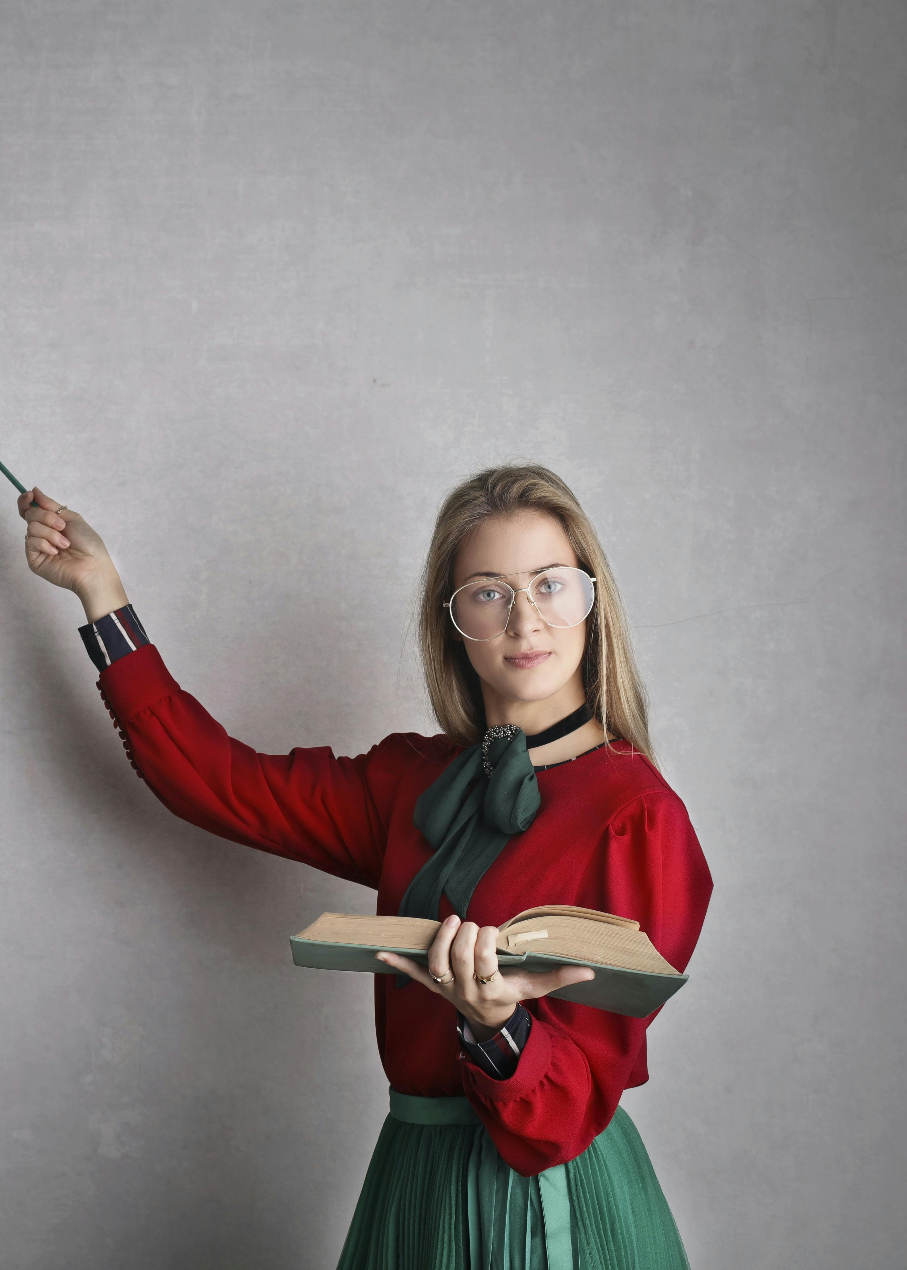 Blonde girl in glasses dressed in red with open book in her left hand pointing with a wand to a specific spot on a blackboard.