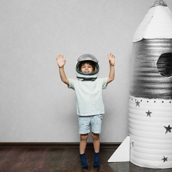 Un bambino con un casco da astronauta esulta a braccia alzate vicino a un razzo di cartone.