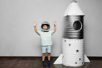 Un bambino con un casco da astronauta esulta a braccia alzate vicino a un razzo di cartone.