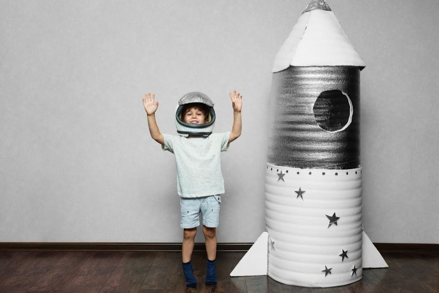 A child in an astronaut helmet exults with arms raised near a cardboard rocket.