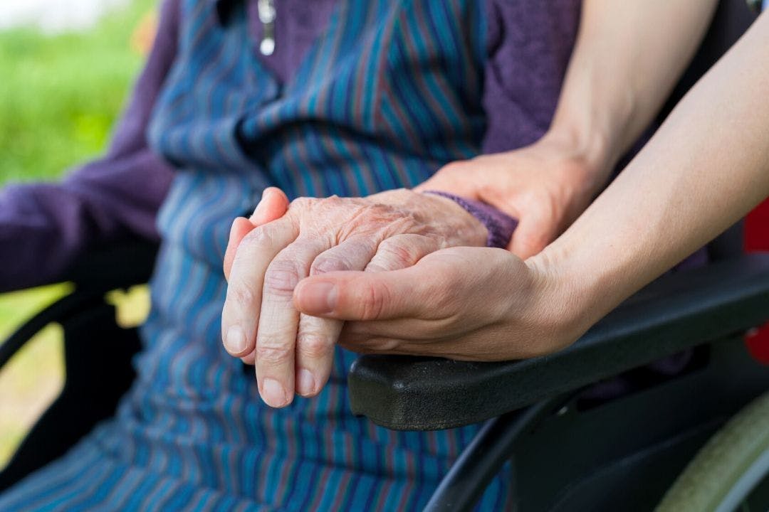 A hand of an elderly person in a wheelchair held by two young hands