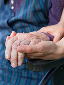 A hand of an elderly person in a wheelchair held by two young hands