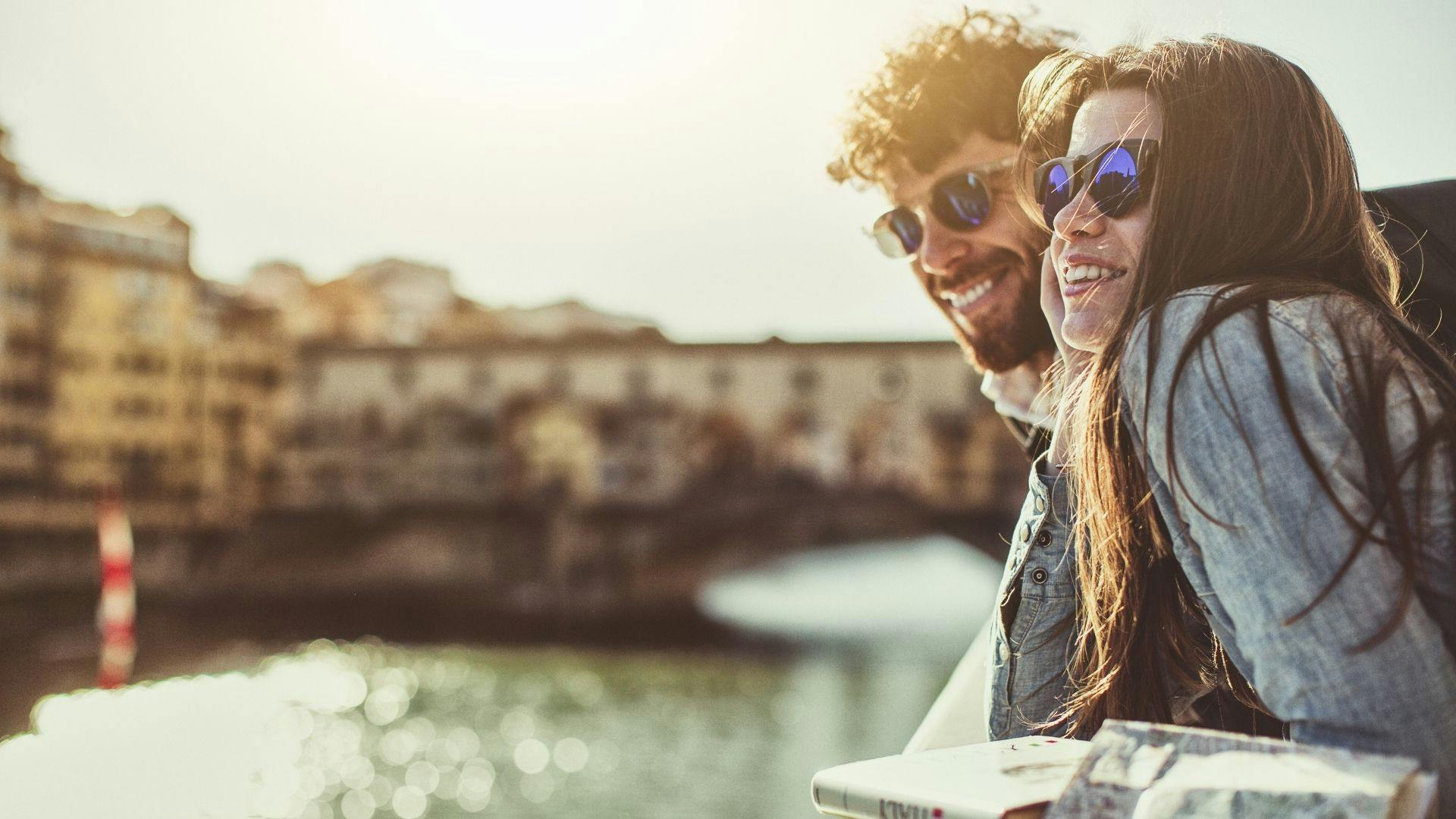 Due ragazzi sorridenti con occhiali da sole guardano il fiume Arno appoggiati al muretto con sullo sfondo il Ponte Vecchio