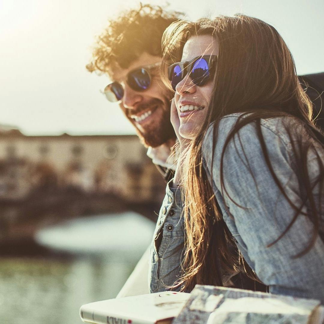 Two smiling guys with sunglasses looking at the Arno River leaning against a low wall with Ponte Vecchio in the background
