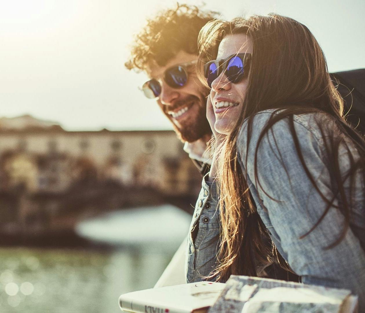 Due ragazzi sorridenti con occhiali da sole guardano il fiume Arno appoggiati al muretto con sullo sfondo il Ponte Vecchio