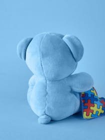Un orsetto di pelouche azzurro di spalle che tiene vicino un cuore di pezza multicolore su sfondo azzurro.