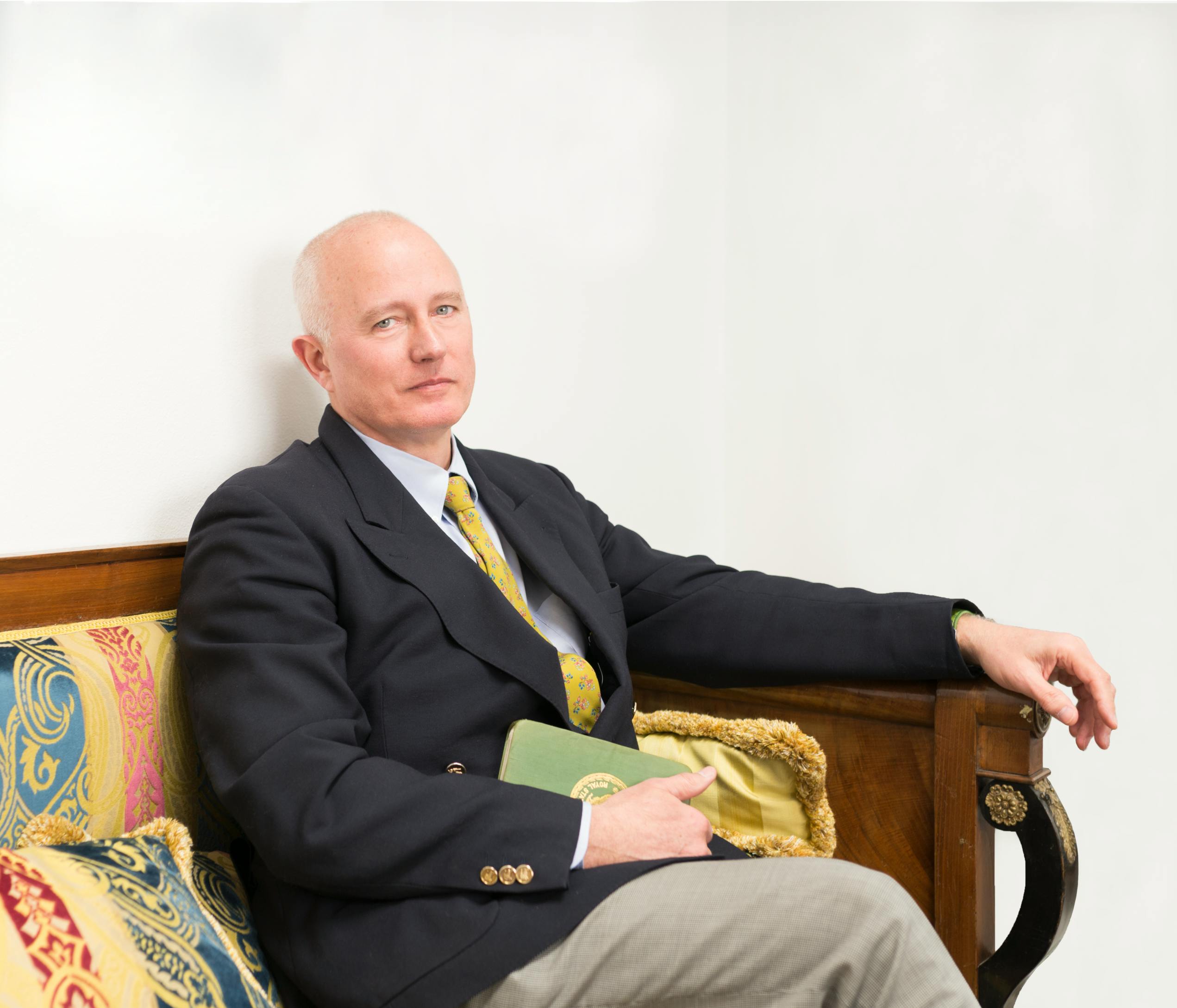 Dr. Stefano Pallanti sitting on an antique sofa with his left arm resting on an armrest and in his right hand a green notebook.