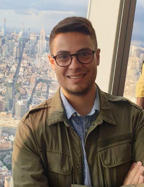 Young man with light brown hair smiling and with his arms crossed. In the background of the picture lies the city of New York.