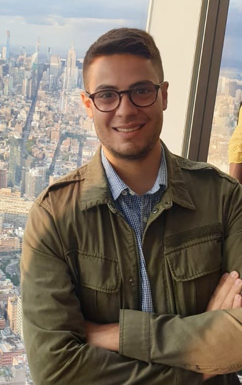 Ragazzo con capelli castani e chiari sorridente e a braccia incrociate. Sullo sfondo della foto la città di New York.