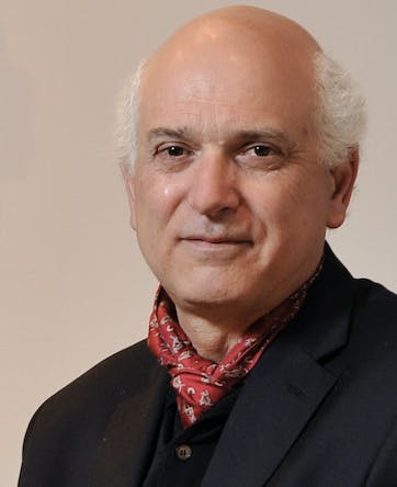 Smiling man with white hair and dark eyes dressed in black suit and red scarf.