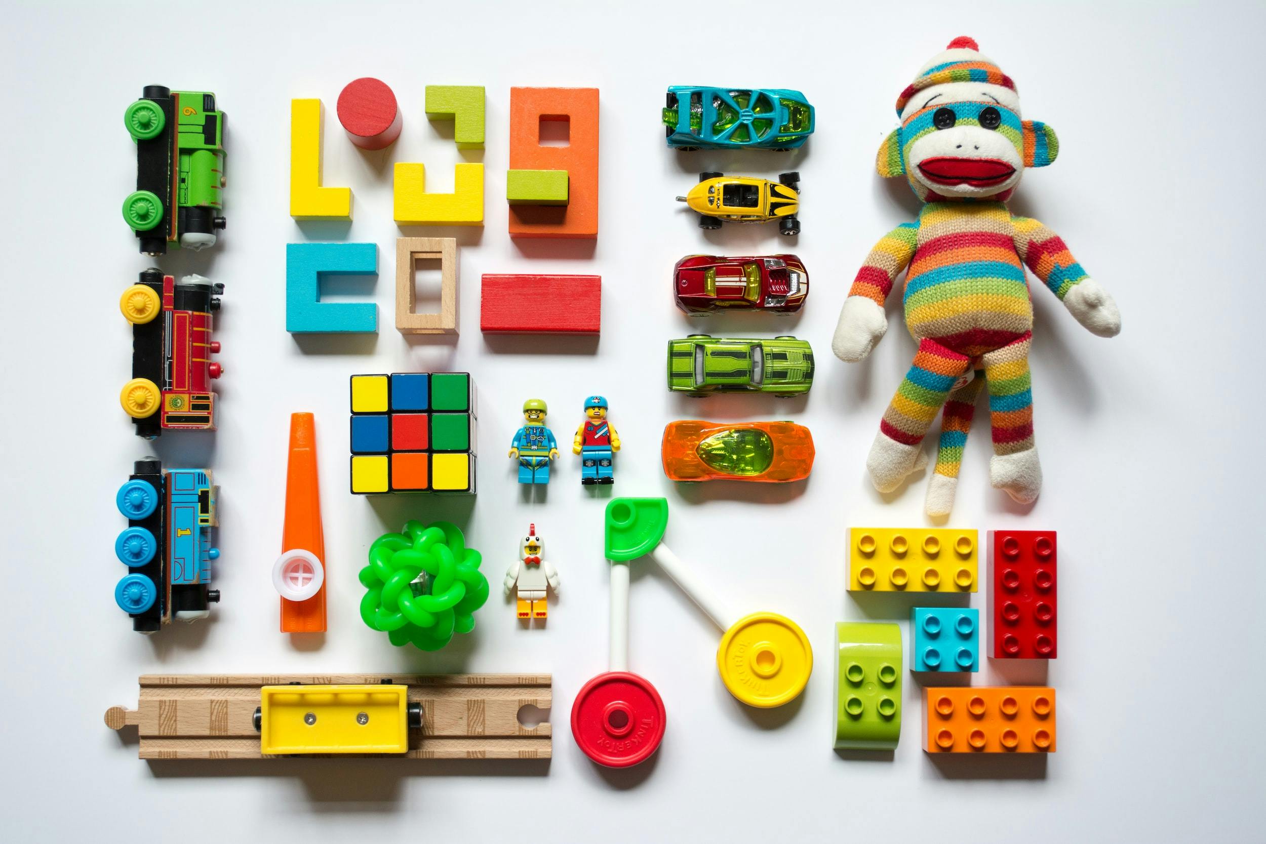 Serie di giocattoli colorati (trenini, rotaie, lego, macchinine, cubo di Rubik, scimmietta di pezza) disposti in griglia in perfetto ordine.