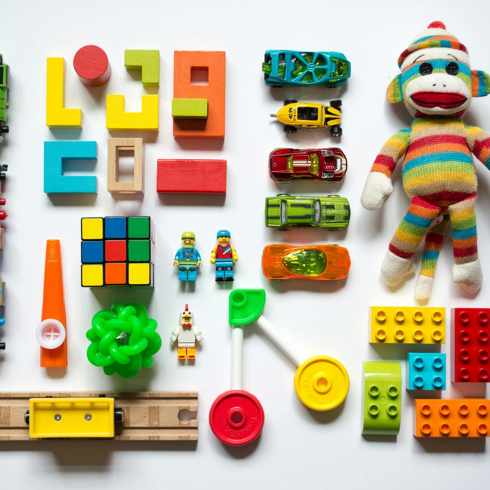 Serie di giocattoli colorati (trenini, rotaie, lego, macchinine, cubo di Rubik, scimmietta di pezza) disposti in griglia in perfetto ordine.
