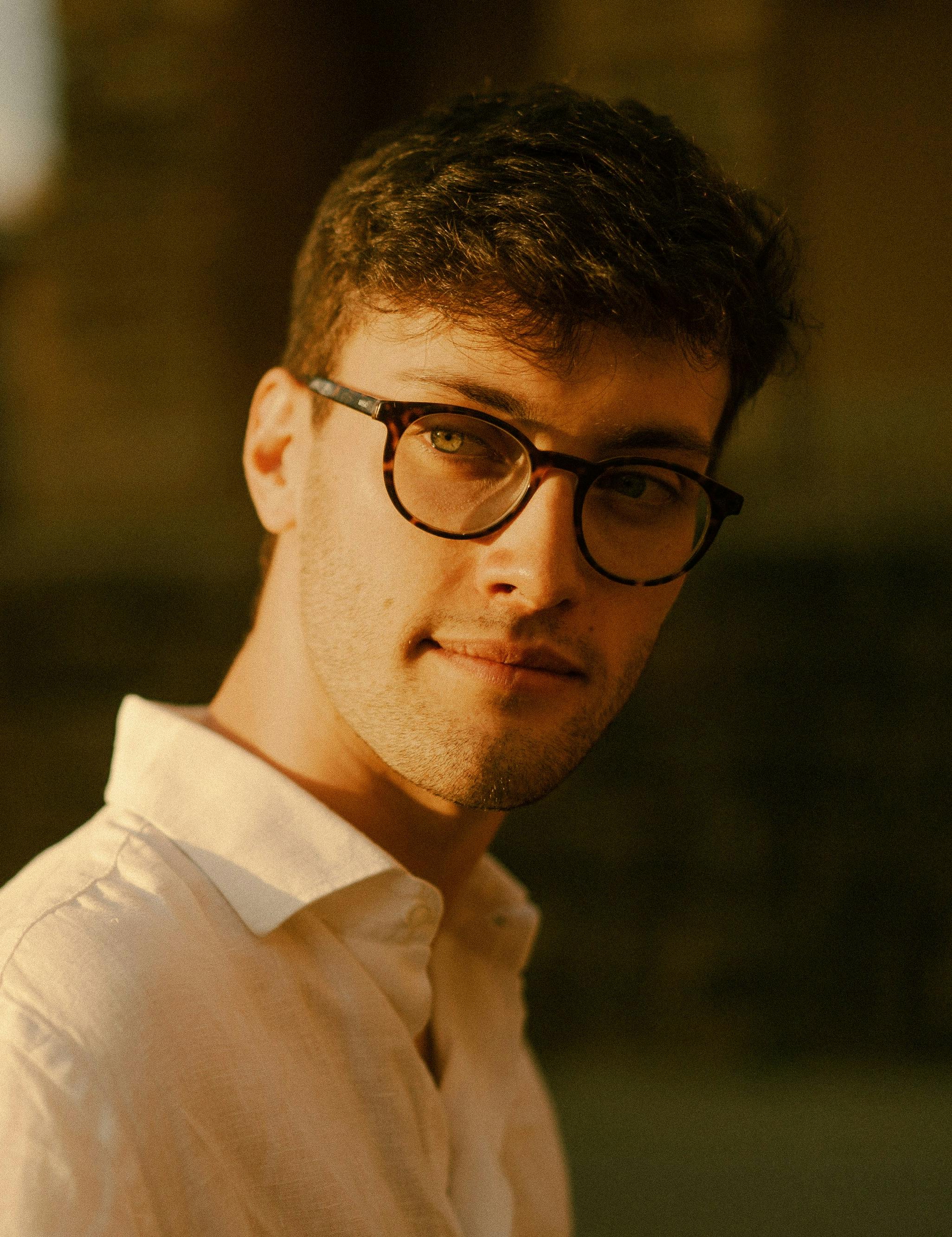 Young man with brown hair, round glasses and green eyes in white shirt, sunlit on the face from the left side.