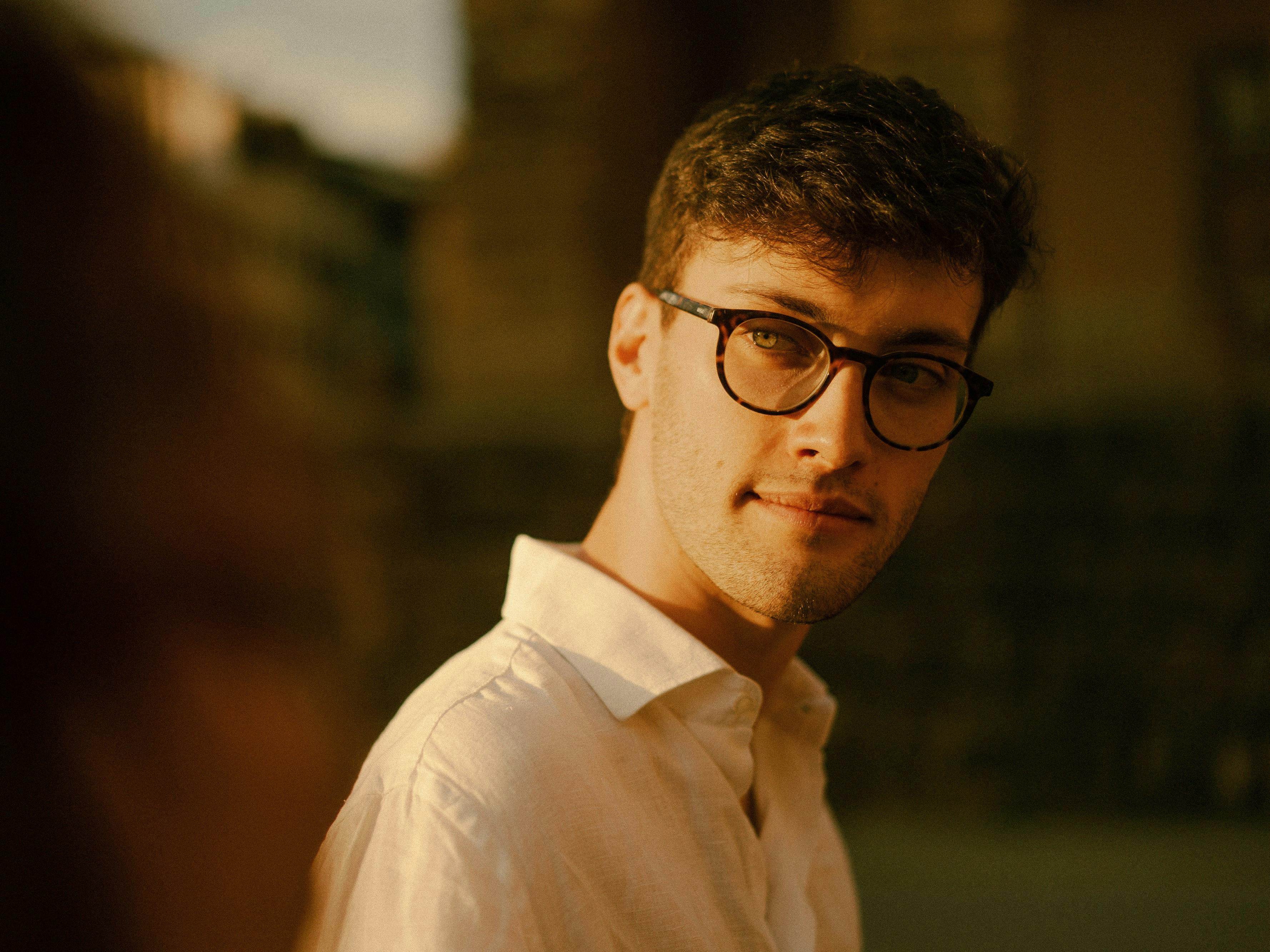 Young man with brown hair, round glasses and green eyes in white shirt, sunlit on the face from the left side.