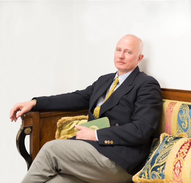Prof. Stefano Pallanti seduto su un divano d'epoca con un braccio appoggiato sul bracciolo di legno e un taccuino verde tenuto nell'altra mano.